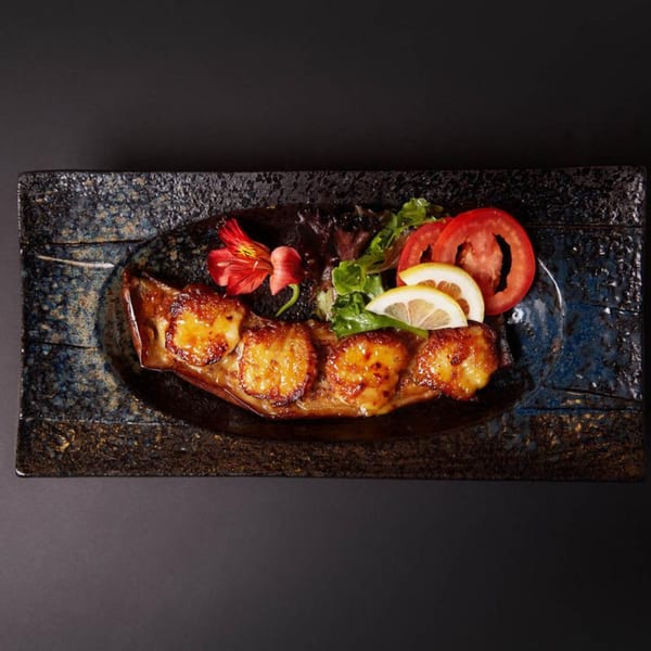 Raku's grilled scallops with lemon, tomato, and greens on a dark, textured plate.
