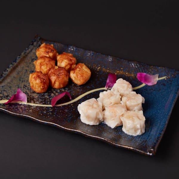 A blue and brown rectangular plate holds fried dumplings and steamed shumai, garnished with purple petals.