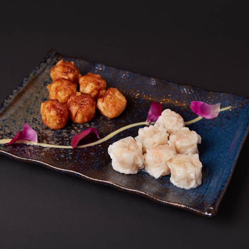 A blue and brown rectangular plate holds fried dumplings and steamed shumai, garnished with purple petals.