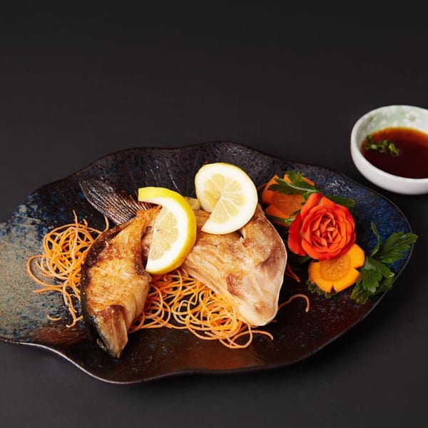 Grilled fish with lemon slices, shredded carrots, and a carved carrot flower on a dark plate.