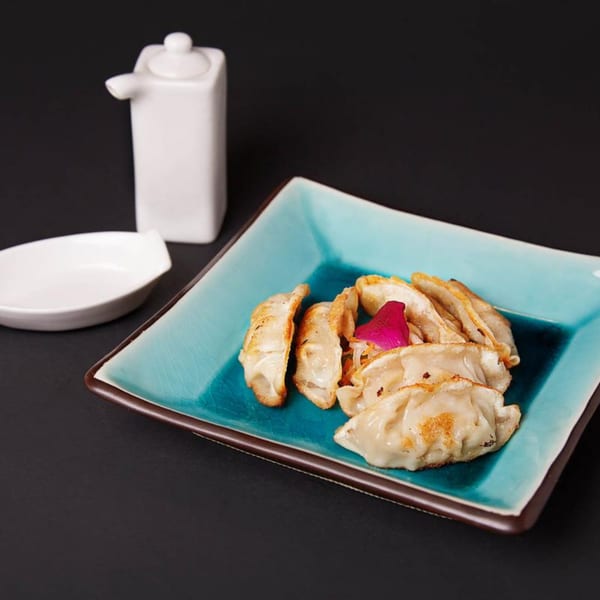 Pan-fried gyoza with a pink garnish on a turquoise plate, with a white sauce dispenser and dish.