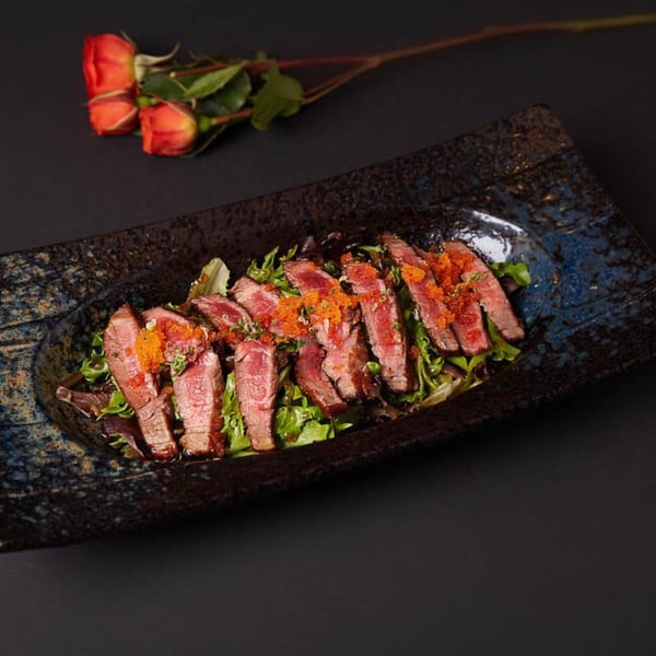 Raku's sliced beef tataki with tobiko and greens on a dark, textured plate, with roses.