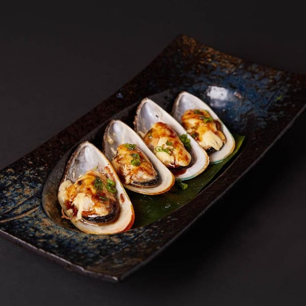 Four baked mussels with sauce and green onions on a dark, textured rectangular plate.